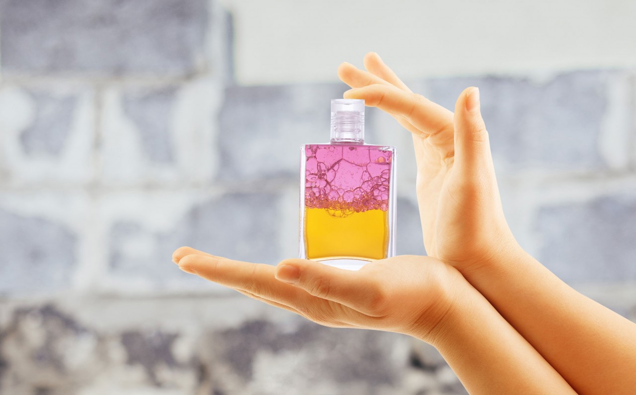 Aura-Soma Equilibrium Anwendung Frauenhände halten eine geschüttelte Aura-Soma-Flasche vor einer strukturierten Wand.