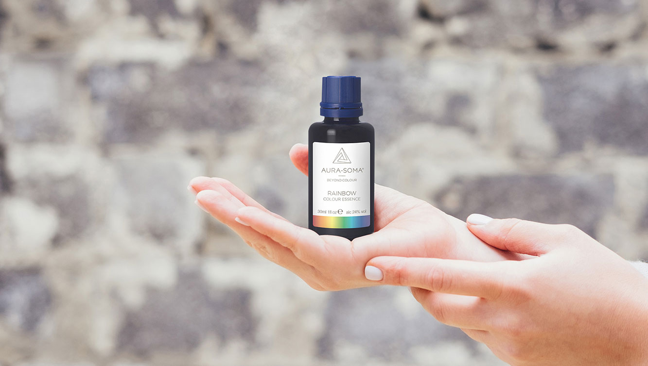 Aura-Soma Banner Farbessenzen Die Aura-Soma Farbessenz Flasche Rainbow in eine rFrauenhand vor einer schönen Steinmauer