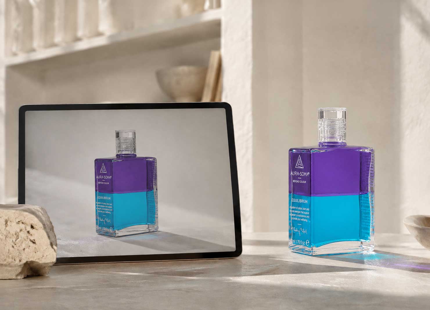 Aura-Soma Equilibrium Flasche B24 in Violett und Türkis neben einem Tablet, das dieselbe Flasche mit leichten Farbabweichungen auf dem Bildschirm zeigt
