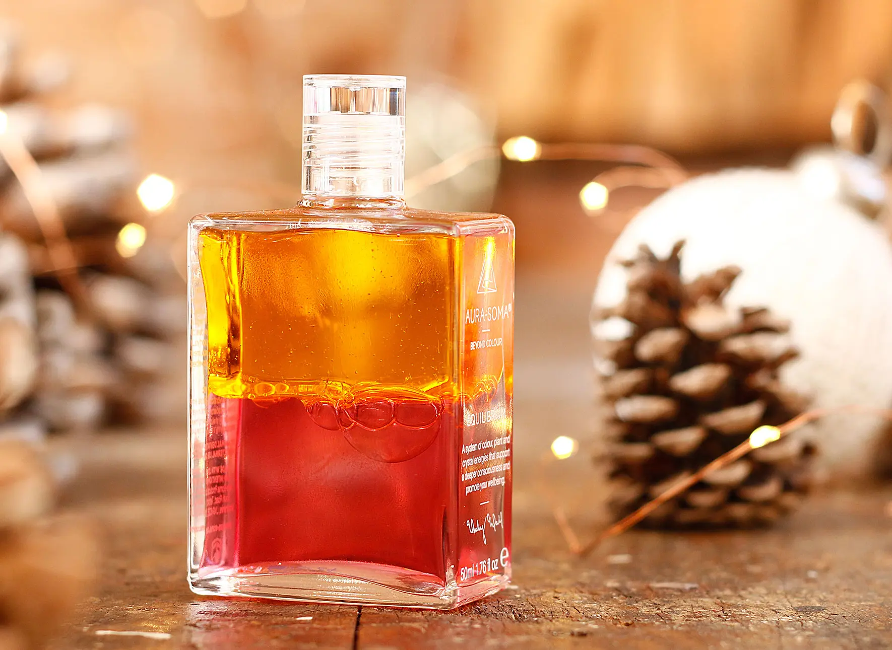 Aura-Soma Equilibrium Weihnachtsbanner Aura-Soma Equilibrium Flasche B115 Erzengel Ariel, in Orange über Rot, weihnachtlich dekoriert mit einem Kiefernzapfen und einer Lichterkette. Die Flasche steht auf einer urigen Holzoberfläche.