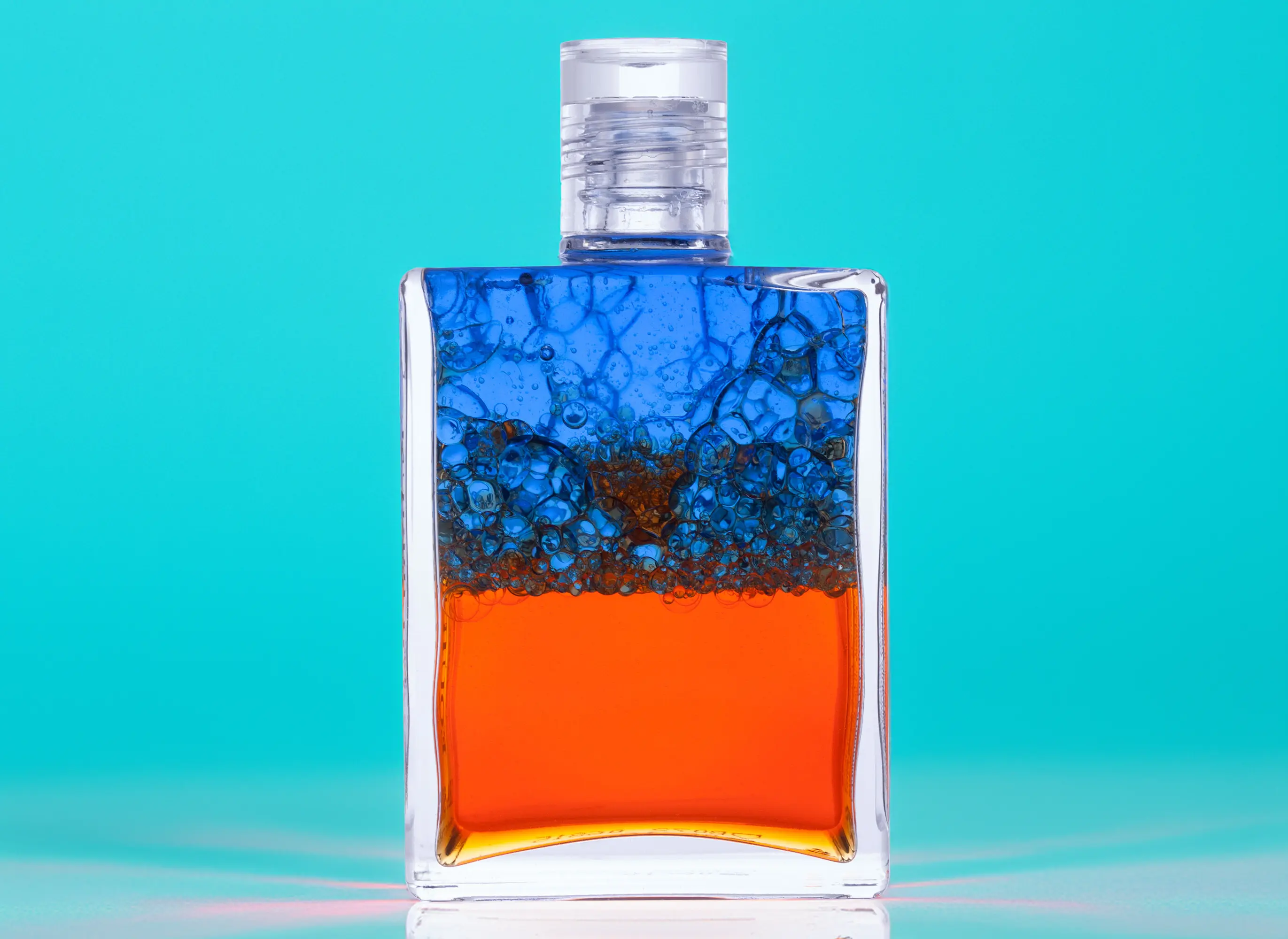 Aura-Soma Farbflasche in Rot-Blau, verschüttelt, vor einem türkisen Hintergrund.