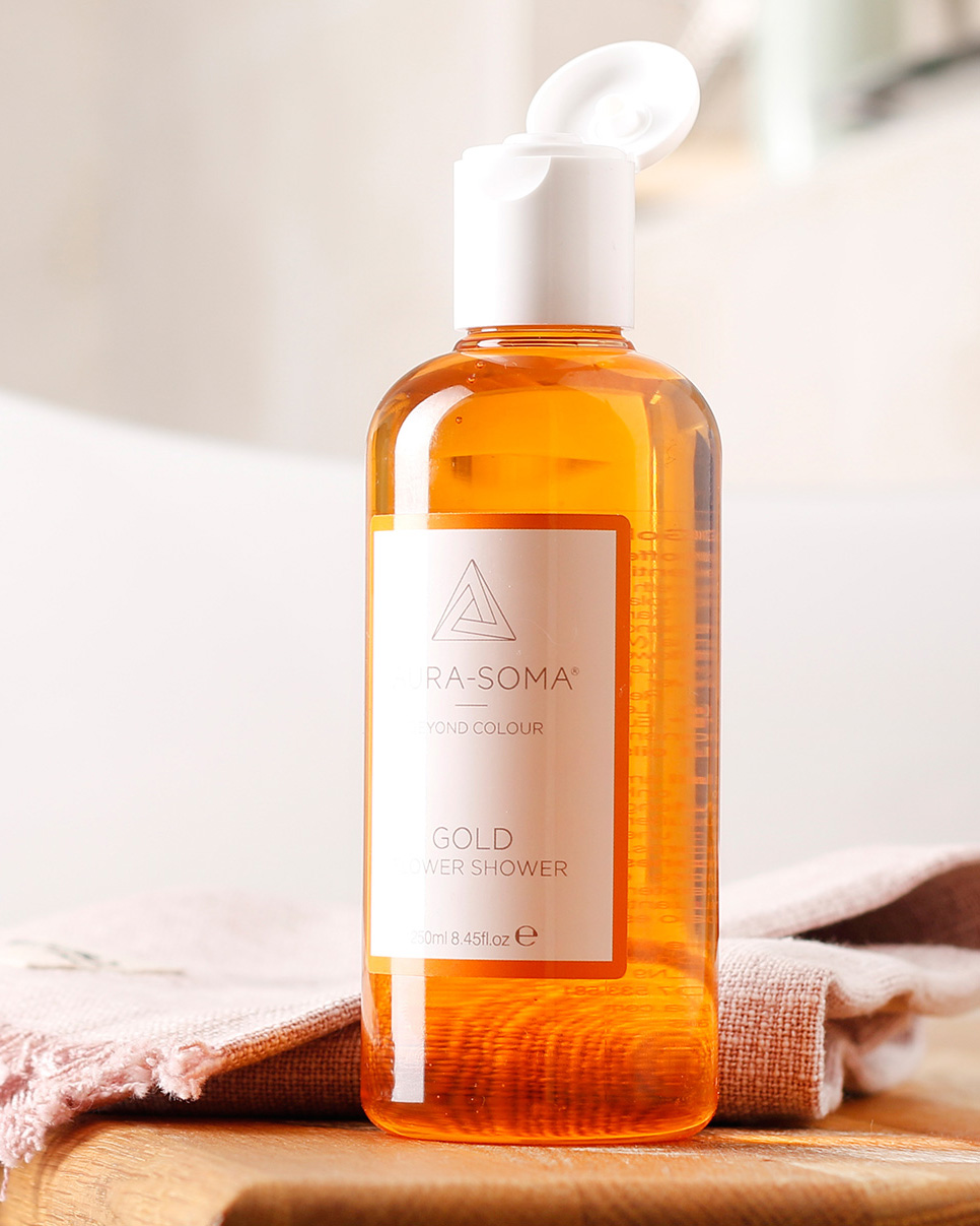 Aura-Soma Flower Shower Gold auf einem hellen Badezimmerhocker neben Handtuch und Dusche